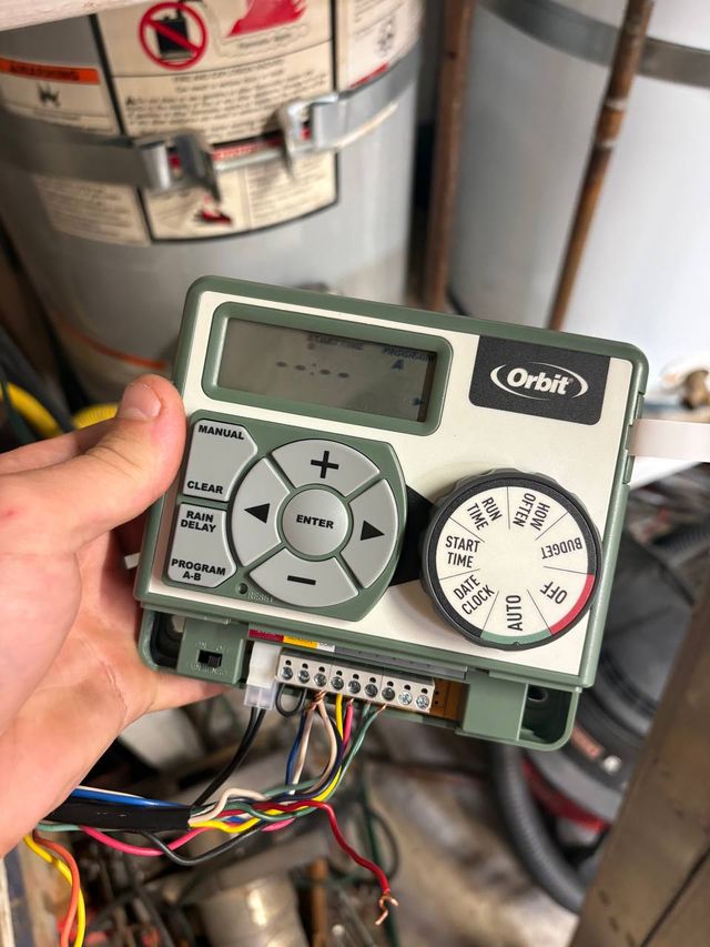 A hand holding an Orbit irrigation controller away from a garage wall, its cover open and the wiring terminal strip exposed.
