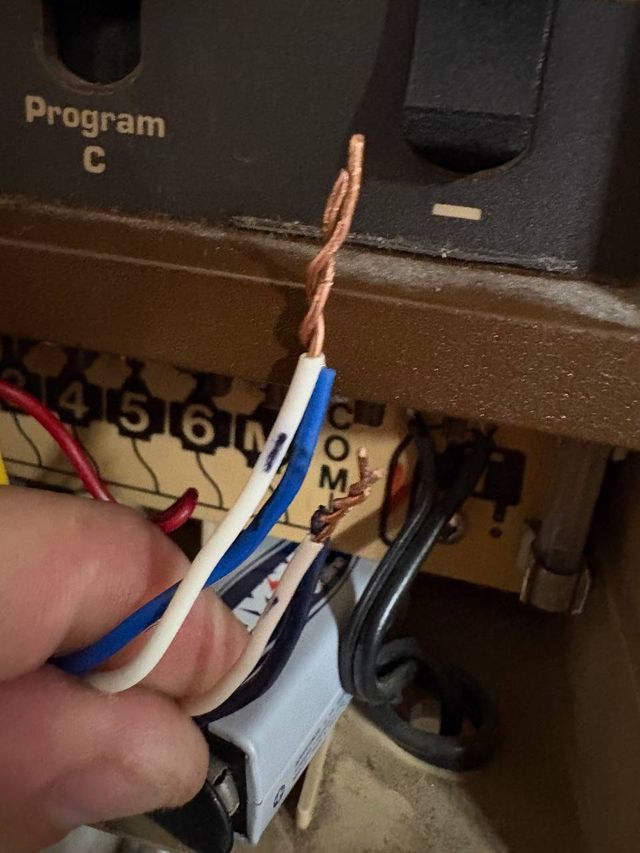 A hand holding hand-twisted copper wire ends in front of a controller terminal strip, with a corroded wire still landed on the board.