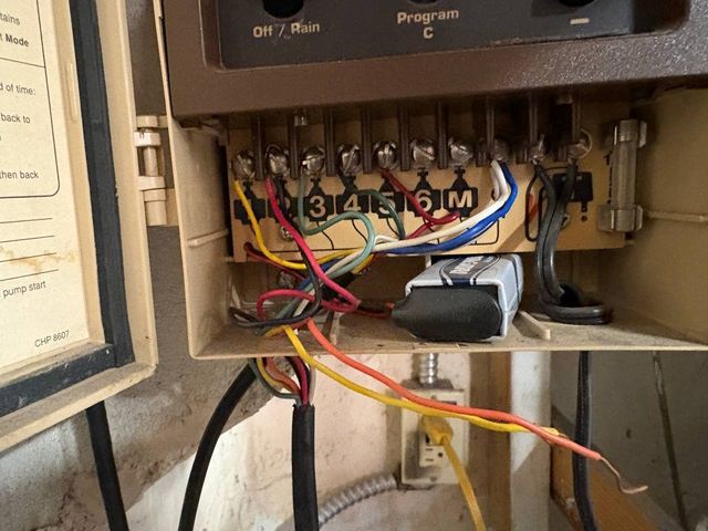 An open older irrigation controller showing a numbered terminal strip with red, yellow, green, white and blue field wires landed on the screw terminals.