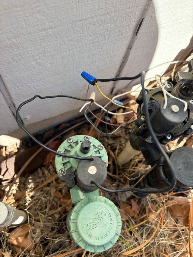 A green Orbit anti-siphon valve and its solenoid standing in dry leaf litter beside a house wall, wire splices capped above it.