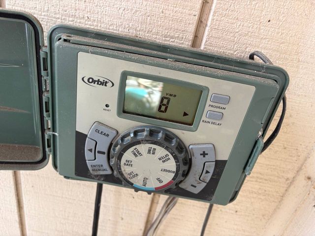 A dusty Orbit irrigation controller mounted on a painted exterior wall with its cover swung open, display showing a single digit.